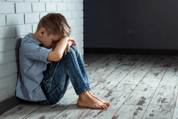 A young boy sits alone with a sad feeling at school near the wall. Offended child abandoned in the corridor and bent against a brick wall. Bullying, discrimination with copy space.