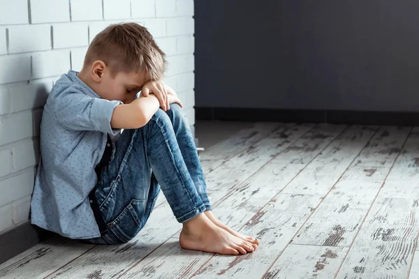 A young boy sits alone with a sad feeling at school near the wall. Offended child abandoned in the corridor and bent against a brick wall. Bullying, discrimination with copy space.