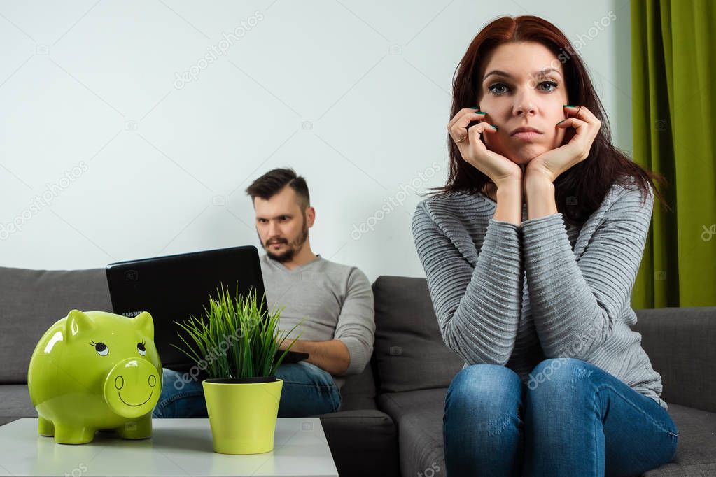 Unhappy wife sits bored, in the background her husband plays on a laptop while sitting on the sofa. The concept of apathy with relationships, boredom, personal space, copy space.