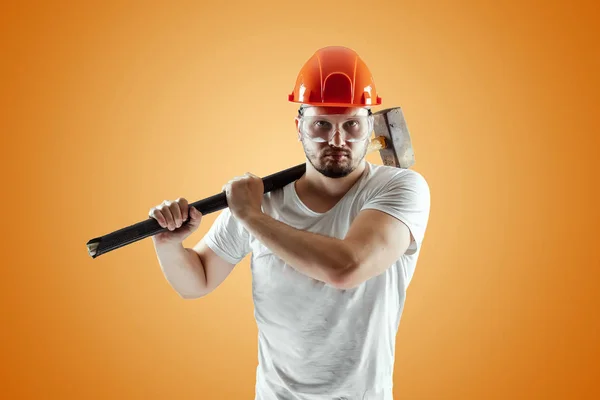 Bearded Man in een helm houdt een Sledgehammer op een oranje achtergrond. Concept bouw, aannemer, herstelling. — Stockfoto