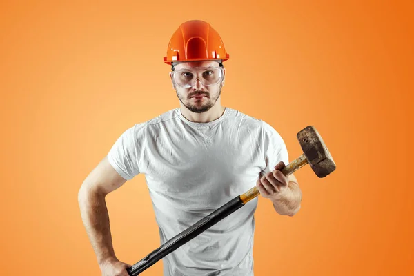 Bearded Man in een helm houdt een Sledgehammer op een oranje achtergrond. Concept bouw, aannemer, herstelling. — Stockfoto