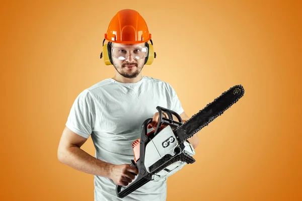 Bearded man in a helmet holds a chainsaw on an orange background. Construction concept, contractor, repair, lumberjack. — Stock Photo, Image