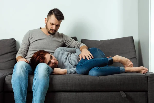 Un hombre lamenta a una chica que tiene dolor de estómago. Siéntate en un sofá gris. Dolor abdominal, ciclo menstrual, problemas de las mujeres . —  Fotos de Stock