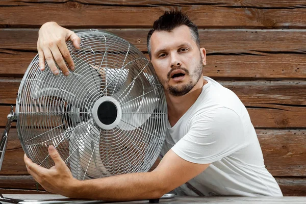 Seorang pria berkeringat memeluk ventilator pendingin, menyegarkan dalam konsep musim panas yang panas. Konsep keselamatan dari panas, cuaca panas, AC primitif — Stok Foto