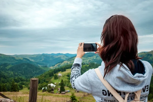 Una joven turista con una mochila de pie en la parte superior de una montaña fotografías en un teléfono un paisaje de montaña. El concepto de vacaciones activas, viajes, vacaciones en las montañas —  Fotos de Stock