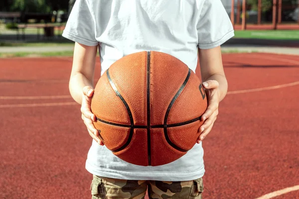 Hält der Junge vor dem Hintergrund eines Basketballfeldes eine Basketballnahaufnahme in den Händen. das Konzept eines sportlichen Lebensstils, Trainings, Sport, Freizeit, Urlaub. — Stockfoto