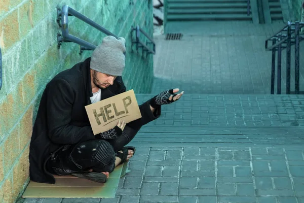 Un hombre, sin hogar, una persona pide limosna en la calle con un cartel de ayuda. Concepto de indigente, adicto, pobreza, desesperación . —  Fotos de Stock