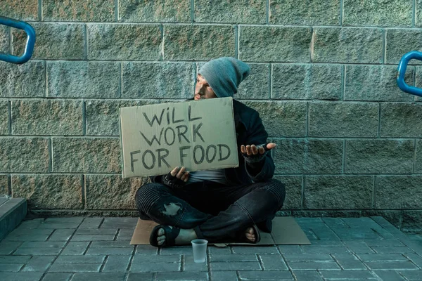 Un uomo, senzatetto, un uomo chiede elemosina per strada con un cartello funzionerà per il cibo. Concetto di senzatetto, tossicodipendente, povertà, disperazione . — Foto Stock