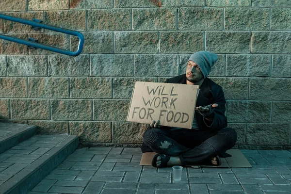 A man, homeless, a man asks for alms on the street with a sign will work for food. Concept of homeless person, addict, poverty, despair. — Stock Photo, Image