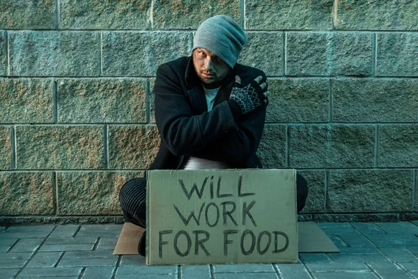 A man, homeless, a man asks for alms on the street with a sign will work for food. Concept of homeless person, addict, poverty, despair. — Stock Photo, Image