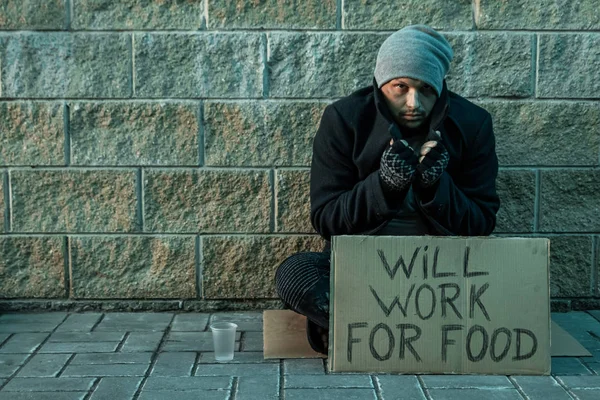 Un homme, SDF, un homme demande l'aumône dans la rue avec une pancarte va travailler pour la nourriture. Concept de sans-abri, toxicomane, pauvreté, désespoir . — Photo