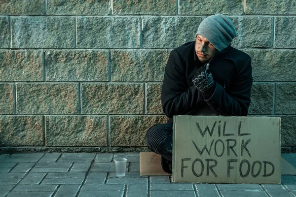 Een man, dakloos, een man vraagt om aalmoes op straat met een bordje zal werken voor eten. Concept van dakloze, verslaafde, armoede, wanhoop. — Stockfoto