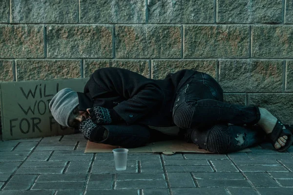 Un hombre, sin hogar, un hombre durmiendo en un piso frío en la calle con un cartel de ayuda. Concepto de indigente, problema social, adicto, pobreza, desesperación . —  Fotos de Stock