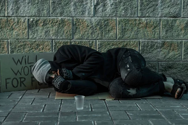 Un homme, sans abri, un homme qui dort sur un sol froid dans la rue avec un panneau d'aide. Concept de sans-abri, problème social, toxicomane, pauvreté, désespoir . — Photo