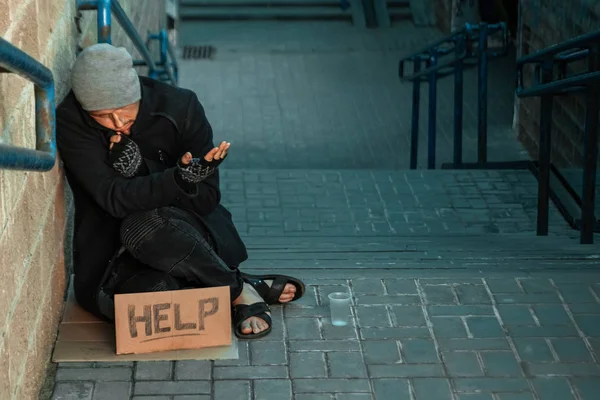 Un hombre, sin hogar, una persona pide limosna en la calle con un cartel de ayuda. Concepto de indigente, adicto, pobreza, desesperación . —  Fotos de Stock