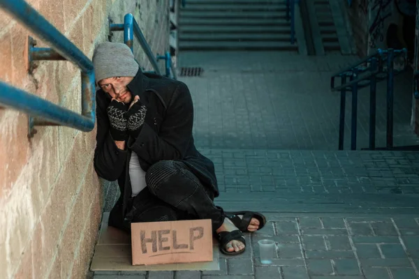 Un uomo, senzatetto, un uomo che dorme su un pavimento freddo per strada con un cartello d'aiuto. Concetto di senzatetto, problema sociale, tossicodipendente, povertà, disperazione . — Foto Stock