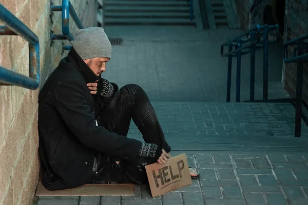 Un hombre, sin hogar, una persona pide limosna en la calle con un cartel de ayuda. Concepto de indigente, adicto, pobreza, desesperación . — Foto de Stock