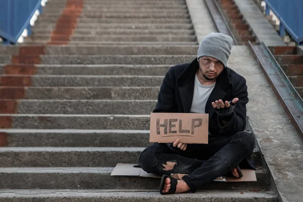 Un hombre, sin hogar, una persona pide limosna en la calle con un cartel de ayuda. Concepto de indigente, adicto, pobreza, desesperación . —  Fotos de Stock