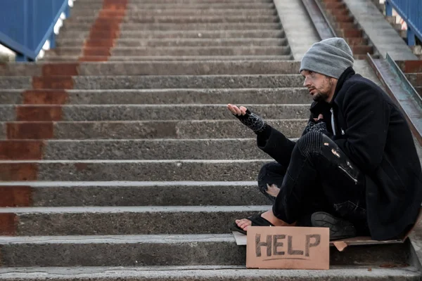 Un hombre, sin hogar, una persona pide limosna en la calle con un cartel de ayuda. Concepto de indigente, adicto, pobreza, desesperación . — Foto de Stock