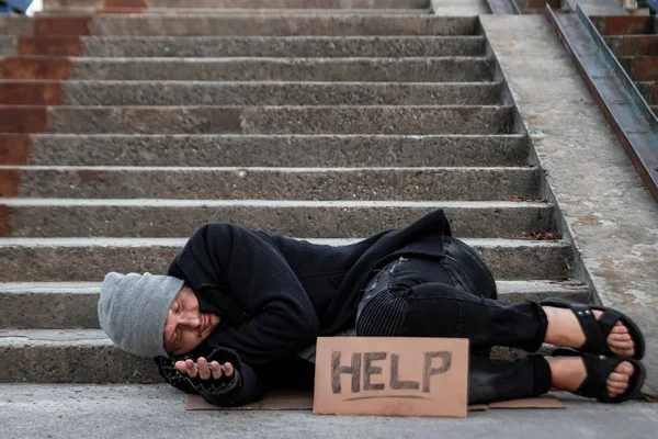 Un uomo, senzatetto, un uomo che dorme su un pavimento freddo per strada con un cartello d'aiuto. Concetto di senzatetto, problema sociale, tossicodipendente, povertà, disperazione . — Foto Stock