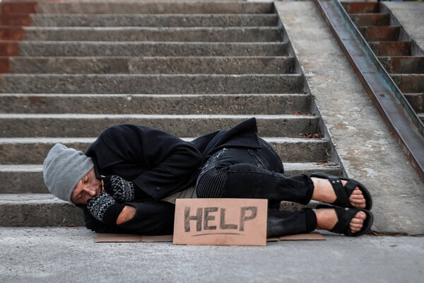 A man, homeless, a man sleeping on a cold floor in the street with a Help sign. Concept of a homeless person, social problem, addict, poverty, despair.
