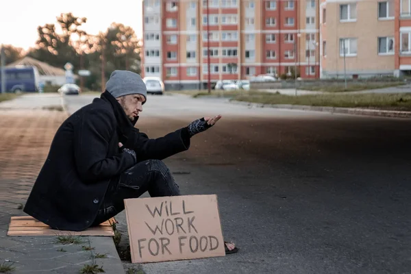 A man, homeless, a man asks for alms on the street with a sign will work for food. Concept of a homeless person, social problem, addict, poverty, despair.