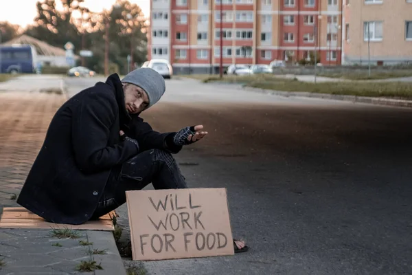 Um homem, sem-teto, um homem pede esmola na rua com um sinal vai trabalhar para a comida. Conceito de um sem-teto, problema social, viciado, pobreza, desespero . — Fotografia de Stock