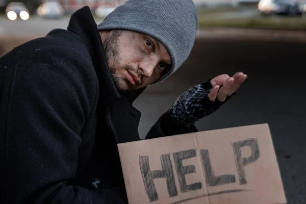 Un uomo, senzatetto, una persona chiede elemosina per strada con un cartello di aiuto. Concetto di senzatetto, tossicodipendente, povertà, disperazione . — Foto Stock