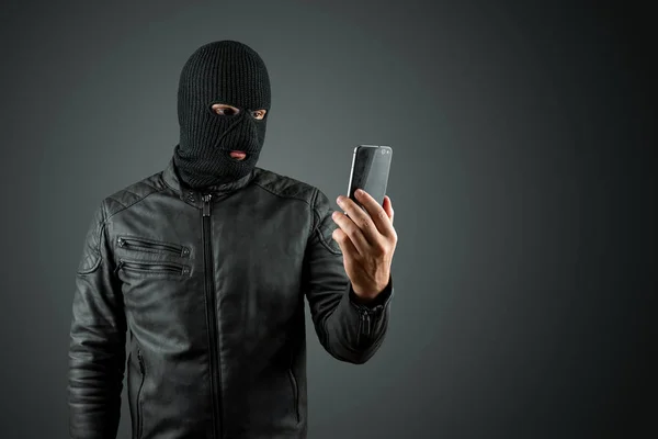 Ladrão, bandido numa balaclava com um telefone nas mãos sobre um fundo preto. Roubo, hacker, crime, roubo. Espaço de cópia — Fotografia de Stock