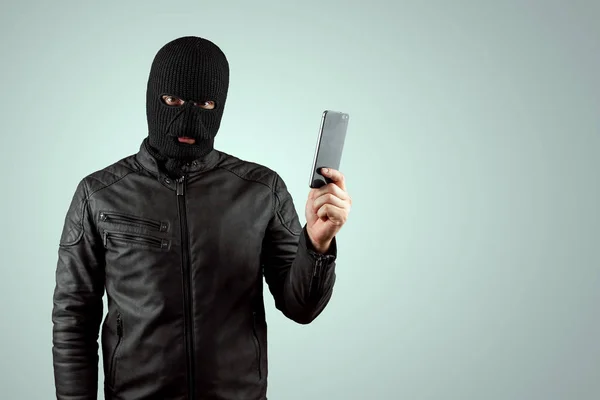 Ladrão, bandido numa balaclava com um telefone nas mãos sobre um fundo claro. Roubo, hacker, crime, roubo. Espaço de cópia . — Fotografia de Stock