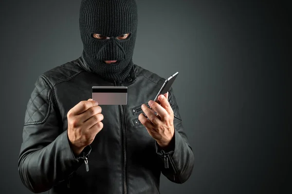 Ladrón, un matón en un pasamontañas tiene una tarjeta de crédito en sus manos sobre un fondo oscuro. Robo, hacker, crimen, robo. Copiar espacio . —  Fotos de Stock