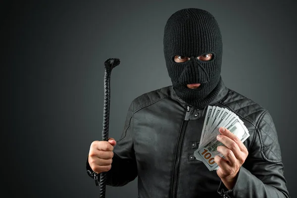 Ladrão, um bandido numa balaclava tem dólares nas mãos num fundo escuro. Roubo, hacker, crime, roubo. Espaço de cópia . — Fotografia de Stock