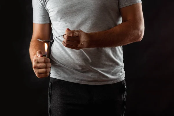 Un drogadicto masculino está preparando drogas en una cuchara con un encendedor en un fondo negro. Problema social, drogadicción, muerte, adicción. Copiar espacio . —  Fotos de Stock