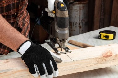 Bir marangoz elektrikli bir yapboz ile tahta bir kirişi keser, erkek eli elektrikli bir yapboz ile yakın plan. Tahtayla çalış.