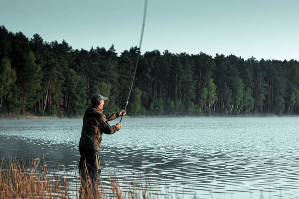 Um pescador masculino no lago está de pé na água e pescando por uma vara de pesca. Conceito de férias hobby de pesca. Espaço de cópia. — Fotografia de Stock