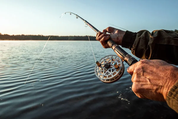 Die Hände eines Mannes in einem Schurkenplan halten eine Angelrute, ein Fischer fängt im Morgengrauen Fische. Angeln Hobby Urlaubskonzept. Kopierraum. — Stockfoto