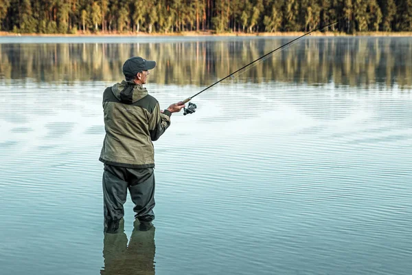 Um pescador masculino no lago está de pé na água e pescando por uma vara de pesca. Conceito de férias hobby de pesca. Espaço de cópia. — Fotografia de Stock