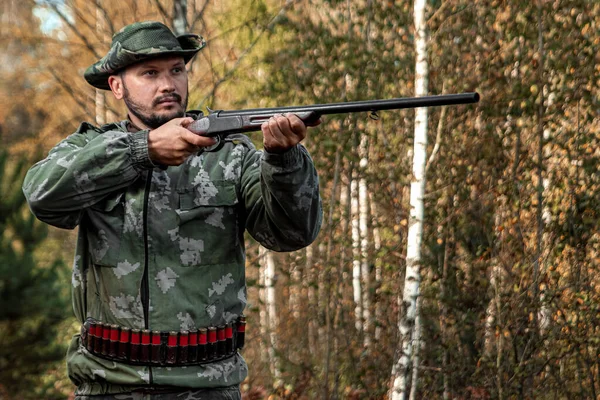 Homem caçador em camuflagem com uma arma durante a caça em busca de pássaros selvagens ou jogo no fundo da floresta de outono. Temporada de caça de outono. O conceito de um hobby, matando . — Fotografia de Stock