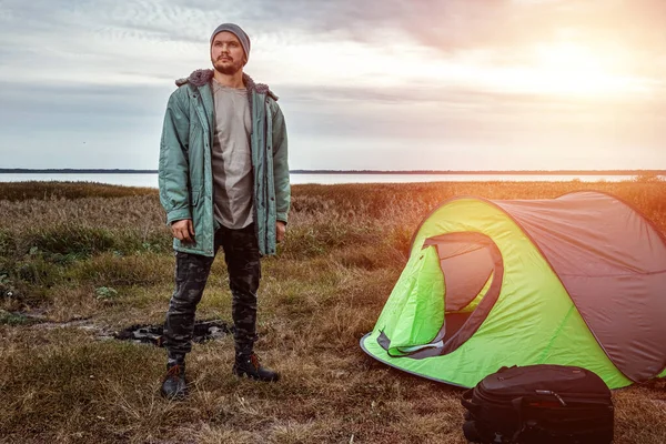 Vousatý muž v zeleném stanu na pozadí přírody a jezera. Koncept cestování, cestovního ruchu, kempování. — Stock fotografie