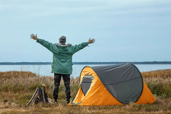 Brodaty mężczyzna w pobliżu namiotu kempingowego w kolorze pomarańczowym na tle natury i jeziora. Koncepcja podróży, turystyki, kempingu. — Zdjęcie stockowe