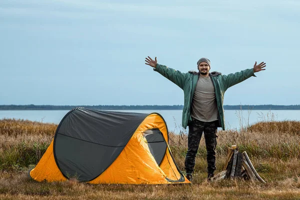 Um homem barbudo perto de uma tenda de acampamento em laranja no fundo da natureza e do lago. O conceito de viagem, turismo, camping . — Fotografia de Stock