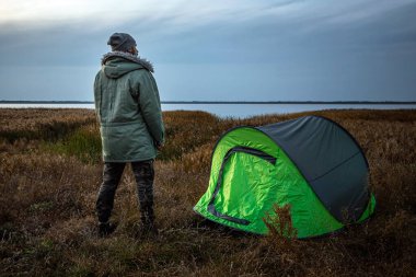 Doğanın ve gölün arka planında yeşil çadırın yanında sakallı bir adam. Seyahat, turizm, kamp konsepti.