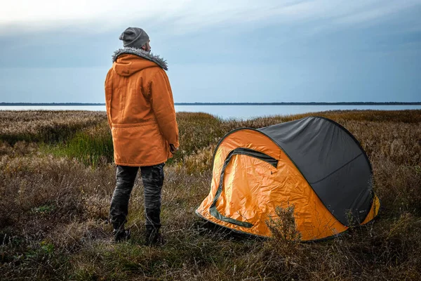 Ein bärtiger Mann in der Nähe eines Zeltes in Orange vor dem Hintergrund der Natur und des Sees. das Konzept von Reisen, Tourismus, Zelten. — Stockfoto