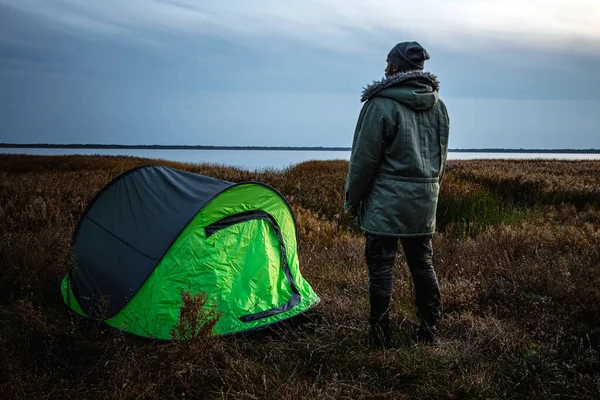 Ein bärtiger Mann in der Nähe eines grünen Zeltes vor dem Hintergrund der Natur und des Sees. das Konzept von Reisen, Tourismus, Zelten. — Stockfoto