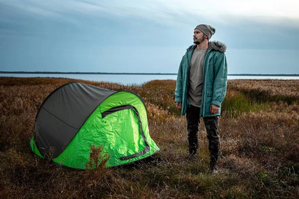 En skäggig man nära ett campingtält i grönt på bakgrunden av naturen och sjön. Begreppet resor, turism, camping. — Stockfoto