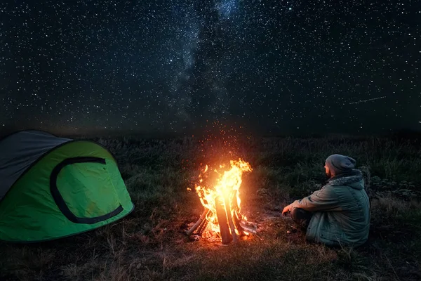 Camping człowiek siedzi przy ognisku w nocy na tle gwiaździstego nieba. Koncepcja podróży, turystyki, kempingu. — Zdjęcie stockowe