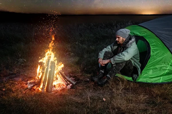 Camping homme assis au coin du feu la nuit contre le ciel. Le concept de voyage, tourisme, camping . — Photo