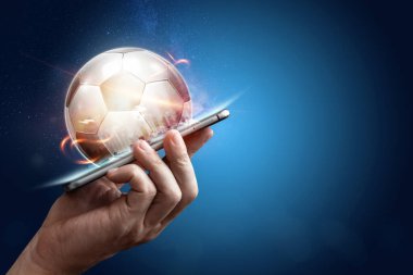 Elinde mavi arka planda 3D futbol topu olan akıllı bir telefon. Bahisler, spor bahisleri, bahisçiler. Karışık ortam