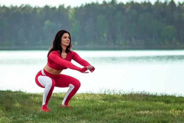 Girl in a sports suit does squats, physical exercises on a background of nature. The concept of a healthy lifestyle, exercise, fresh air. Copy space