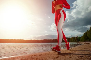 Eşofmanlı bir kızın bacakları yakın plan, göl ya da denizin yolu boyunca koşuyor. Sağlıklı yaşam tarzı konsepti, kardiyo çalışması. Boşluğu kopyala